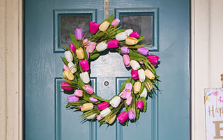 A photograph of a hand made spring wreath for a front door. The spring wreath features artifical tulips in pink, yellow and white. The door of the building is blue which gives the image a fresh new look.