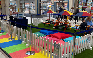 Inside Spinning Gate shopping centre, there are Warm White Festoon Lights hung above of some colourful picnic benches and brightly coloured bean bags that children are sitting on colouring and reading books.