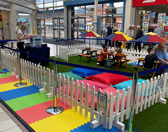 Inside Spinning Gate shopping centre, there are Warm White Festoon Lights hung above of some colourful picnic benches and brightly coloured bean bags that children are sitting on colouring and reading books.